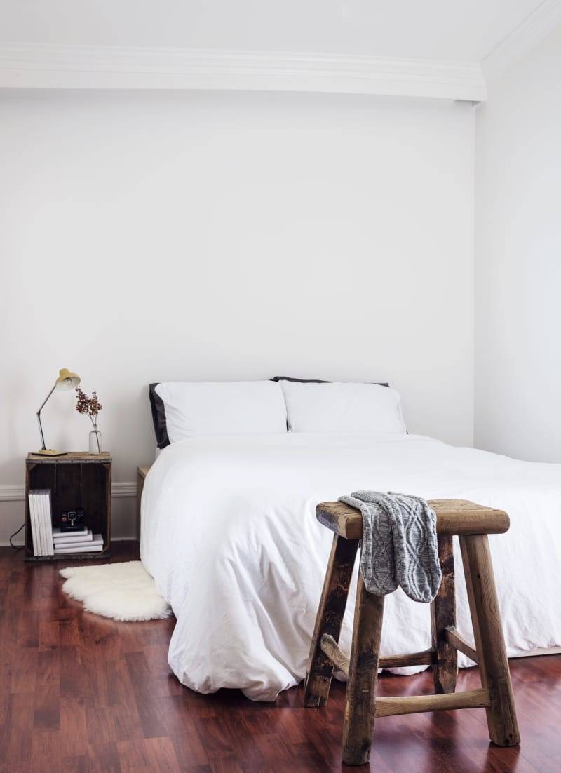 Habitación con pared blanca, suelo de parqué, taburete de madera y mesita de noche con una lámpara